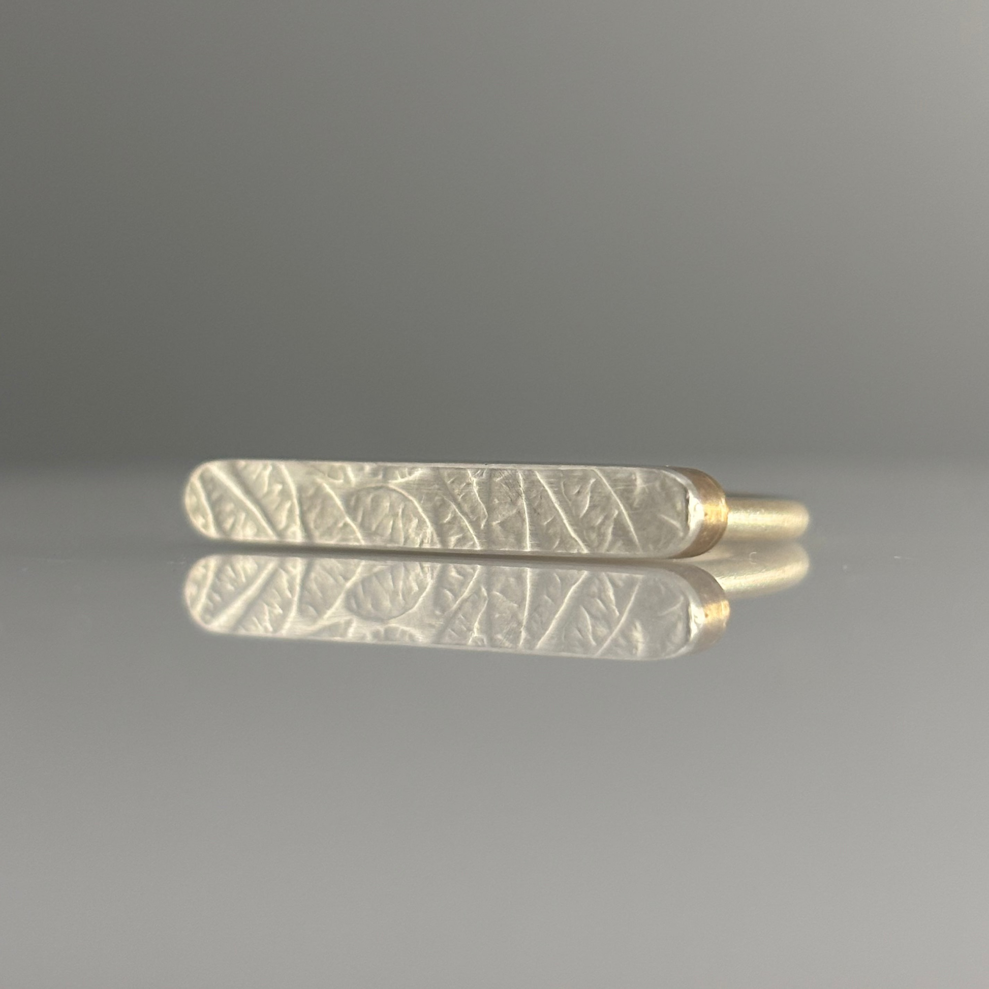 A gold-toned ring featuring a textured, patterned white inlay, displayed horizontally on a reflective gray surface against a neutral gray background.