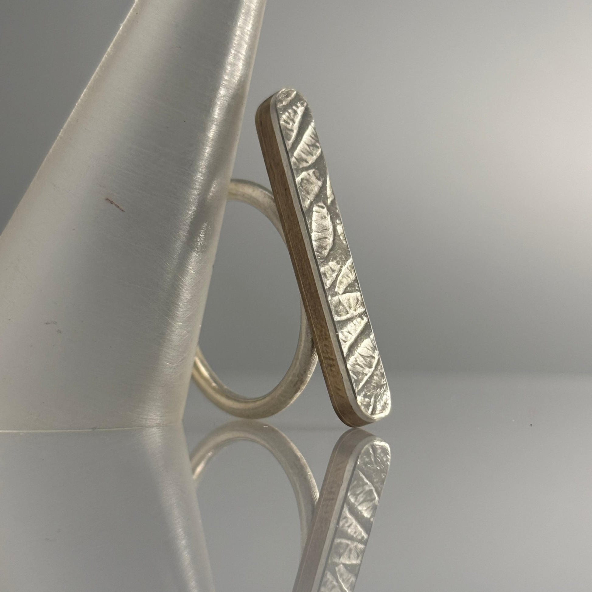 A silver ring with a rectangular, textured face and a simple circular band, displayed on a reflective surface with a frosted cone prop against a gray gradient background.